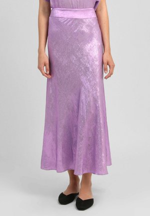 Femme portant une jupe midi lavande brillante et des chaussures noires sans lacets, debout devant un fond gris clair uni.