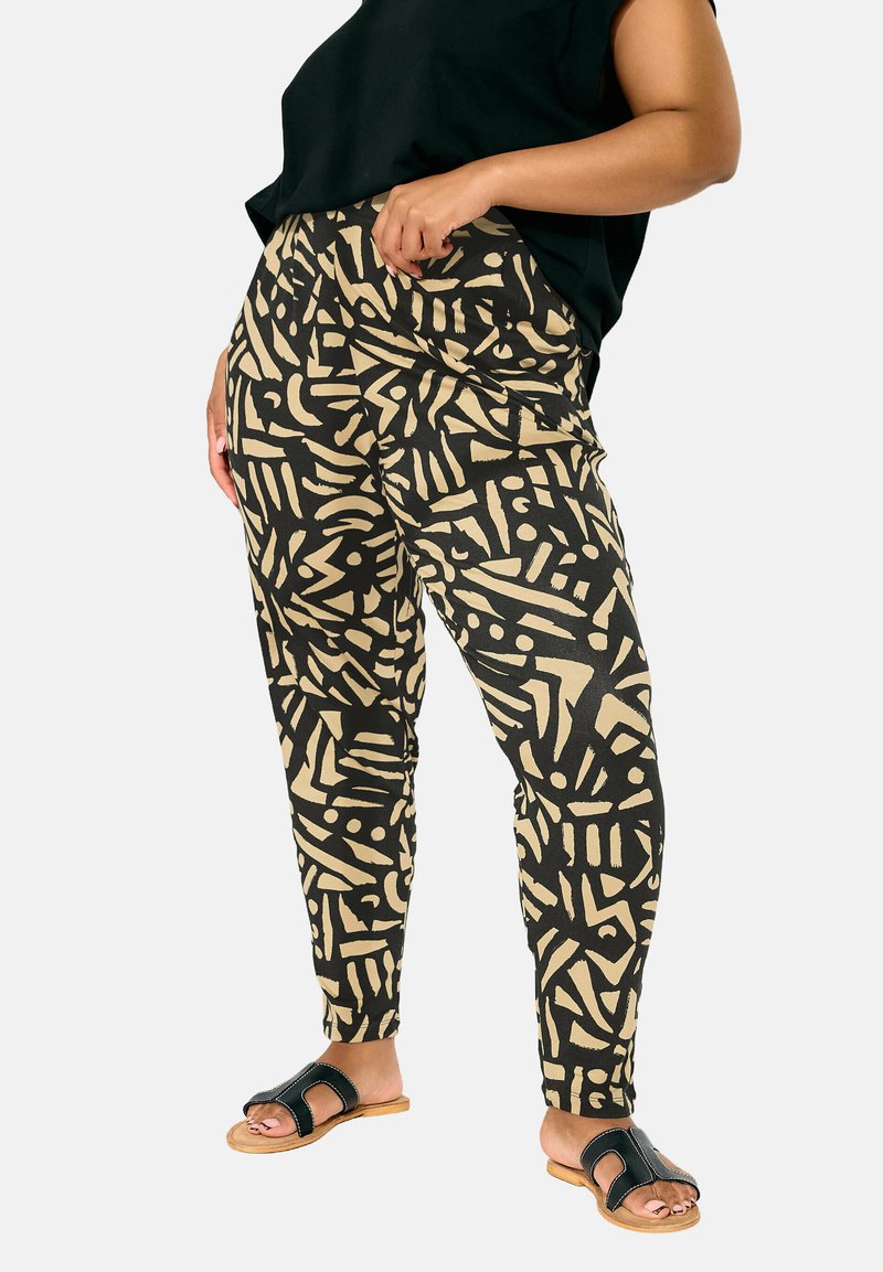 Pantalons noirs et beiges à motifs avec des formes abstraites, fabriqués en tissu doux. Coupe ajustée avec une taille élastique. Portés avec des sandales noires à lanières.