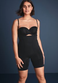 Musta muotoileva bodysuit, jossa on rakenteelliset kupit, säädettävät hihnat ja puolipitkä helma. Suloinen kangas, jossa on saumaton muotoilu ja leikkausdetalji.