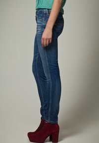 Hubené modré džíny s vybledlým povrchem, prošívanými detaily a pěti kapsami, doplněné červenými semišovými kotníkovými botami na vysokém podpatku.