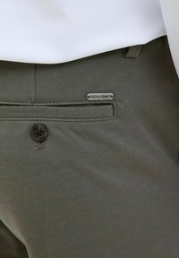 Tamsiai alyvuogių žali kelnės, pagamintos iš audinio, turi priekinį kišenę su juodu mygtuku ir sidabrinę etiketę su "JACK & JONES" ženklu.