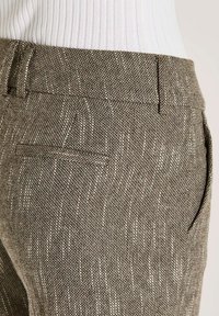 Close-up van gemêleerde bruine tweed broek met riemlussen en een gespleten achterzak, gedragen met een witte geribbelde top.