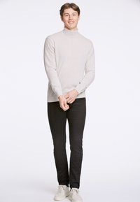 Maglione a collo alto beige chiaro realizzato in un tessuto morbido, abbinato a pantaloni neri aderenti e sneakers bianche, in piedi su uno sfondo neutro.