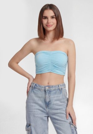Junge Frau mit glatten braunen Haaren, die ein hellblaues gerafftes Tubetop und hellblaue Denim-Cargohosen trägt, posiert mit einer Hand in der Hüfte.