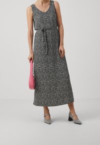 Femme portant une robe midi sans manches noire avec un imprimé floral blanc, tenant un sac à main rose et des chaussures à talons carrés grises, se tenant sur un sol gris.