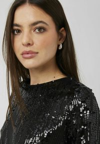Haut à sequins noir avec col haut, présentant une surface texturée de sequins brillants de différentes tailles. De simples boucles d'oreilles en argent complètent le look.