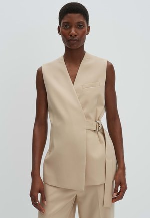Frau trägt ärmellose, beiger, maßgeschneiderter Weste mit D-Ring-Gürtel und passender Hose mit weitem Bein vor einfarbigem Hintergrund.