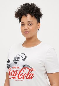 Fehér pamut póló, fekete-fehér Mikulás grafikával, piros "Igyál Coca-Colát" felirattal és sima textúrával.