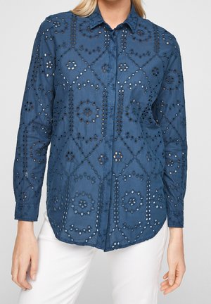Vrouw draagt een langemouwen blauw overhemd met bloemige opengewerkte borduursels en een witte broek tegen een effen achtergrond.