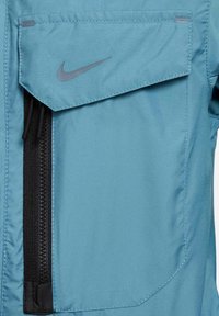 Chaqueta sintética azul con un bolsillo en el pecho con solapa, cremallera lateral y un sutil logo negro de Nike. Textura suave y diseño entallado.