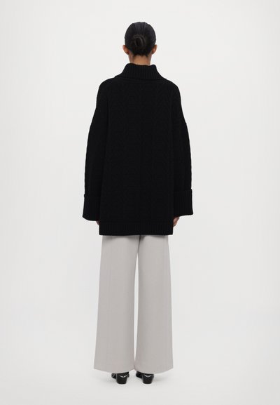 Black cable-knit oversized sweater with a high collar, ribbed cuffs, and hem, paired with pale gray wide-leg trousers.