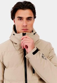 Chaqueta acolchada beige con cuello alto y capucha ajustable. Presenta un cierre frontal con cremallera, acentos negros y un logo en la manga.
