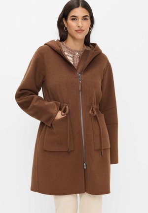 Donna che indossa un cappotto marrone con cappuccio, cerniera frontale e grandi tasche, pantaloni beige e un maglione a fantasia sotto.