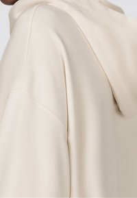 Sudadera beige con capucha, fabricada en tejido suave y liso, con un corte relajado y hombros caídos, que destaca por sus mínimos detalles de costura.