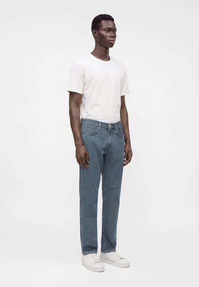Homme debout portant un t-shirt blanc uni, un jean bleu droit et des baskets blanches sur un fond clair uni.