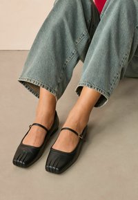 Black leather ballerina flats with a square toe, featuring a strap across the foot and a unique toe design. Worn with cropped light blue denim.