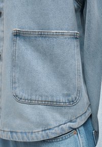 Φωτεινό γαλάζιο τζιν τζάκετ με τσέπη και αντίθετη καφέ ραφή, παρουσιασμένο σε άτομο που φοράει τζιν παντελόνι στο ίδιο ύφος.