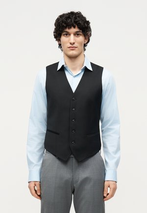 Ung mann med krøllete hår iført lyseblå skjorte, svart knappet vest og grå bukser, stående foran en ensfarget hvit bakgrunn.