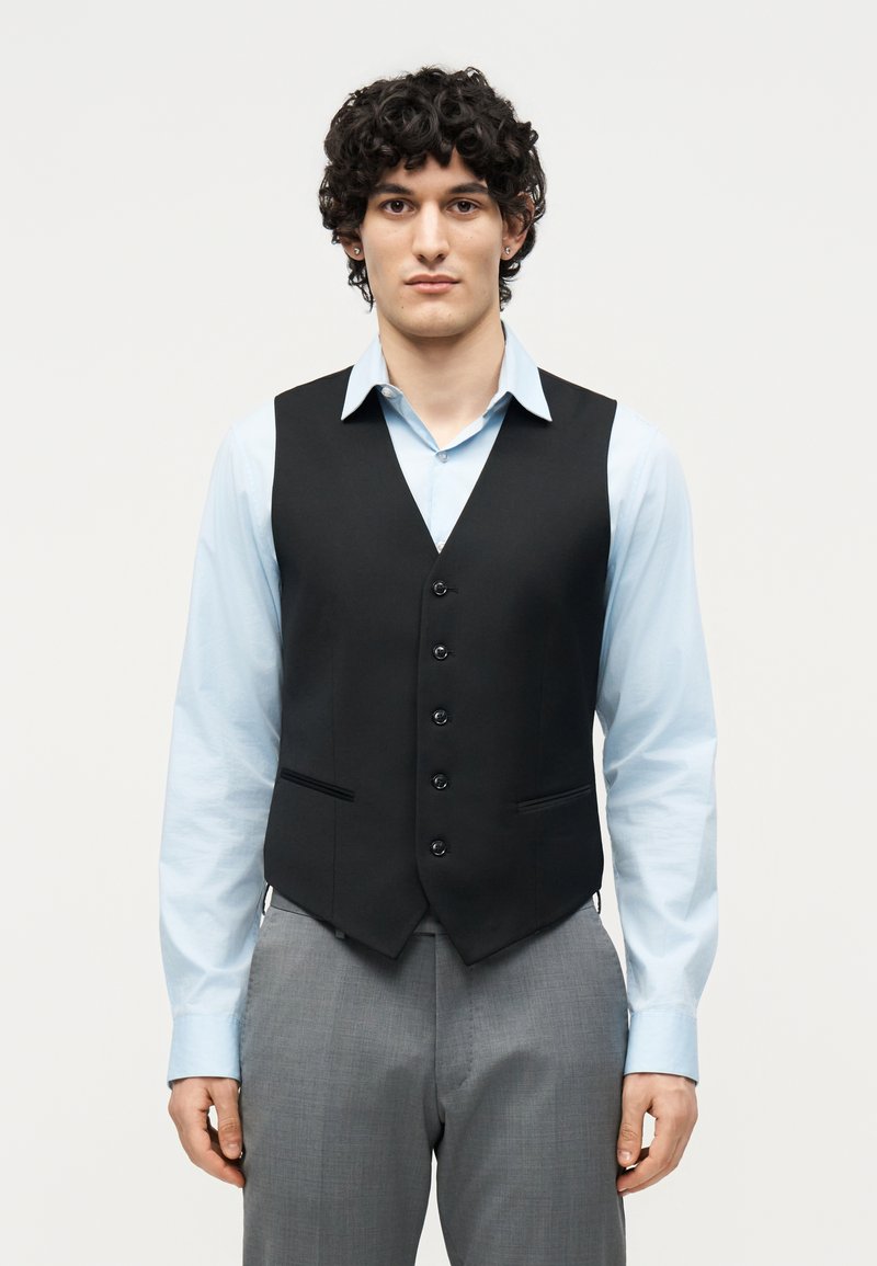 Νέος άνδρας με σγουρά μαλλιά που φοράει ανοιχτό μπλε πουκάμισο, μαύρο γιλέκο με κουμπιά και γκρι παντελόνι, στέκεται μπροστά σε ένα απλό λευκό φόντο.