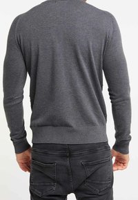 Homme portant un pull gris foncé ajusté à manches longues et un jean noir, vu de dos sur un fond uni.