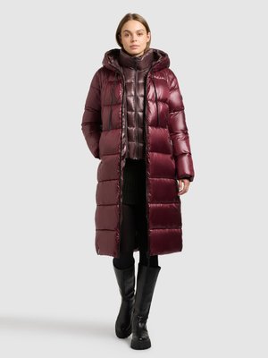 Lange, bordeauxrode gewatteerde jas met capuchon, voorzien van een glanzende textuur en zijzakken. Draagt over een mouwloze, doorgestikte binnenlaag en zwarte laarzen.