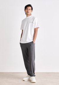 T-shirt bianca a maniche corte con testo nero, abbinata a pantaloni grigi su misura e sneakers bianche. Design semplice e moderno, texture del tessuto liscia.