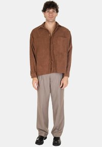 Camicia di velluto a coste marrone con maniche lunghe e taschino sul petto, abbinata a pantaloni grigi con pieghe e scarpe nere. Sfondo bianco.