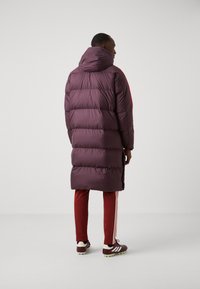 Abrigo puffer burdeos con capucha, con acolchado horizontal. Combinado con pantalones deportivos rojos con franjas blancas y zapatillas deportivas oscuras.