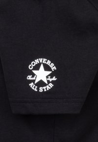 Camisa de tela negra con un logo circular blanco que presenta una estrella, con las palabras "CONVERSE ALL STAR" y "Chuck Taylor" en una tipografía estilizada.