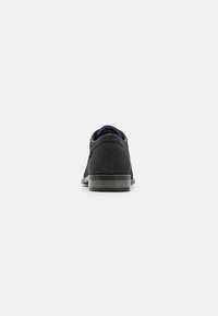 Zapatilla negra con parte superior de malla texturizada, cordones planos, cuello acolchado y suela de goma vista desde atrás.
