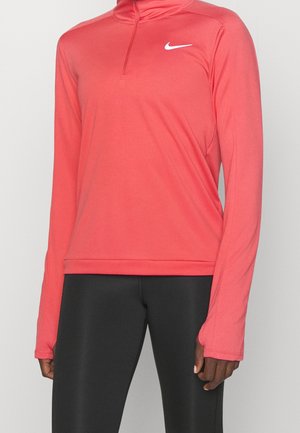 Person trägt ein korallenfarbenes, langärmeliges Sportoberteil mit Viertelreißverschluss und schwarze Leggings und steht vor einem schlichten Hintergrund.
