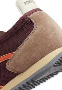 Zapatilla con una parte superior de malla burdeos, acentos de ante marrón y detalles en naranja. Presenta una suela de goma beige y una pestaña en el talón blanca con texto.