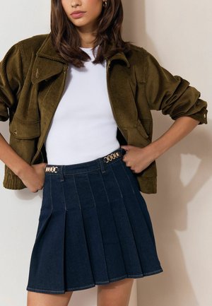 Frau trägt eine olivgrüne Cordjacke, weißes Top und einen dunkelblauen, plissierten Rock mit goldenen Kettendetails, posiert mit Händen in der Hüfte.