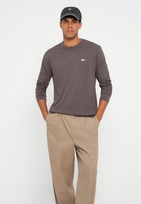 Camicia marrone a maniche lunghe con collo a giro, arricchita da un piccolo logo, abbinata a pantaloni beige plissettati. Il tessuto sembra morbido e informale. Cappellino nero in testa.