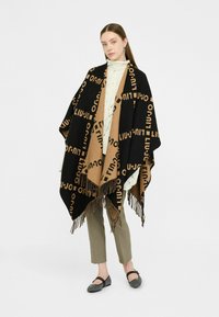 Zwart en tan poncho met "LIU JO" logo patroon, franjes aan de randen, laag over een crèmekleurige trui en gedragen met grijze broek en zwarte schoenen.