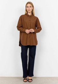 Brown button-up shirt with a collar, featuring long sleeves and a curved hem, paired with dark blue flared jeans and strappy black sandals.