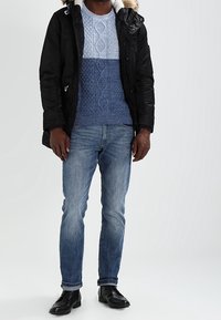 Black parka with fur trim, blue cable-knit sweater, and light blue patterns, paired with medium wash denim and black leather shoes.