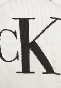 Gestrickter Pullover in Off-White mit auffälligen schwarzen "CK"-Buchstaben. Gerippte Struktur, figurbetontes Design und kontrastreiches Farbschema.