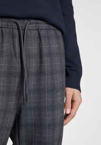 Grå rutiga sweatpants med elastisk midja och dragsko. Texturen är slät och har ett ljust rutig mönster med mörka detaljer.