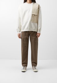 Sudadera blanca con un parche beige y un bolsillo en la parte frontal, combinada con pantalones cargo marrones que tienen bolsillos laterales y detalles de cordón.