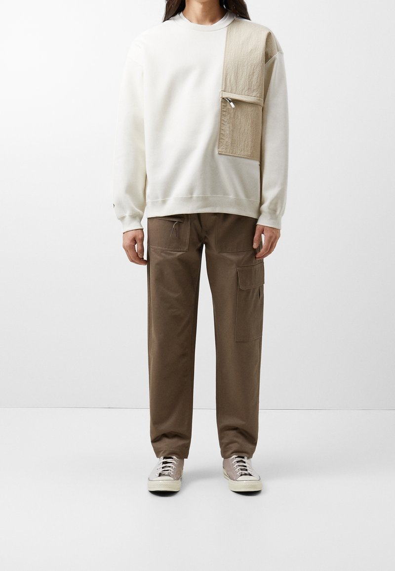 Sudadera blanca con un parche beige y un bolsillo en la parte frontal, combinada con pantalones cargo marrones que tienen bolsillos laterales y detalles de cordón.