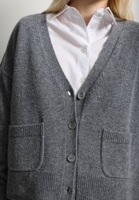 Cárdigan de punto gris con escote en V, dos bolsillos delanteros y cierre de botones. Cuenta con puños y dobladillo acanalados. Lleva puesto un camisa blanca con collar.