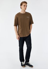 Brun bomulds T-shirt med en afslappet pasform, korte ærmer og et lille hvidt logo på brystet. Sammen med mørk denim jeans og sneakers.