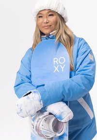 Blå snowboard med "ROXY" logga, som hålls av en person i en matchande blå vinterjacka. Utrustad med vita, molnmönstrade vantar och snowboardbindningar.