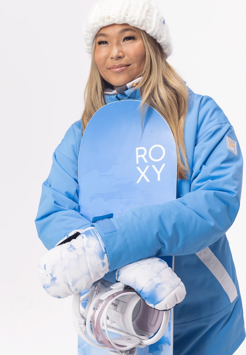 Blå snowboard med "ROXY" logga, som hålls av en person i en matchande blå vinterjacka. Utrustad med vita, molnmönstrade vantar och snowboardbindningar.