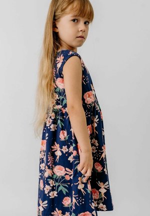 Young girl with long blonde hair wearing a navy blue dress with pink floral and fairy patterns, standing against a plain background.
