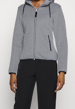 Mujer con una chaqueta con capucha y cremallera con un patrón geométrico en blanco y negro y pantalones negros, de pie con los brazos relajados.