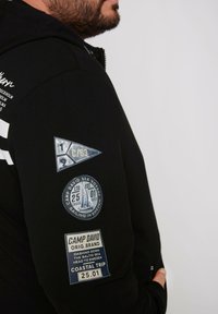 Sudadera con capucha negra hecha de tela suave, que presenta varios parches azules con diseños náuticos y texto en la manga izquierda.