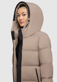Beige péřová bunda s velkou kapucí, černými akcenty a zipovým zapínáním. Má hladkou texturu a prošívaný design pro izolaci.