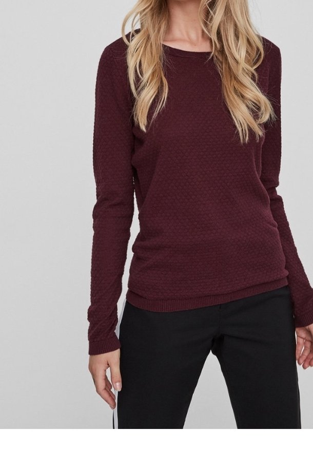 Femme portant un pull bordeaux texturé à manches longues et un pantalon noir, debout devant un fond gris clair uni.